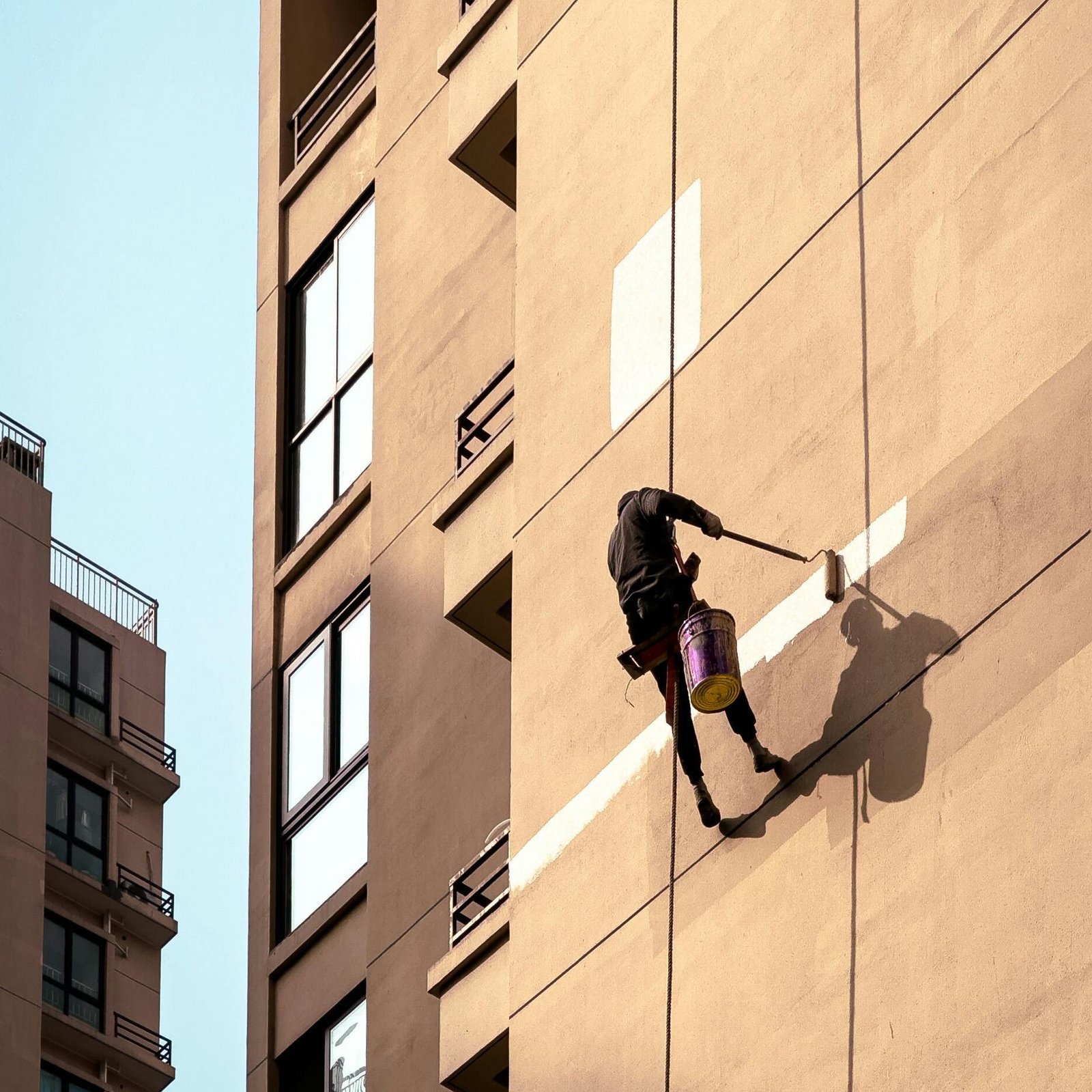 Man painting tall building exterior while suspended outdoors, showcasing urban maintenance work.