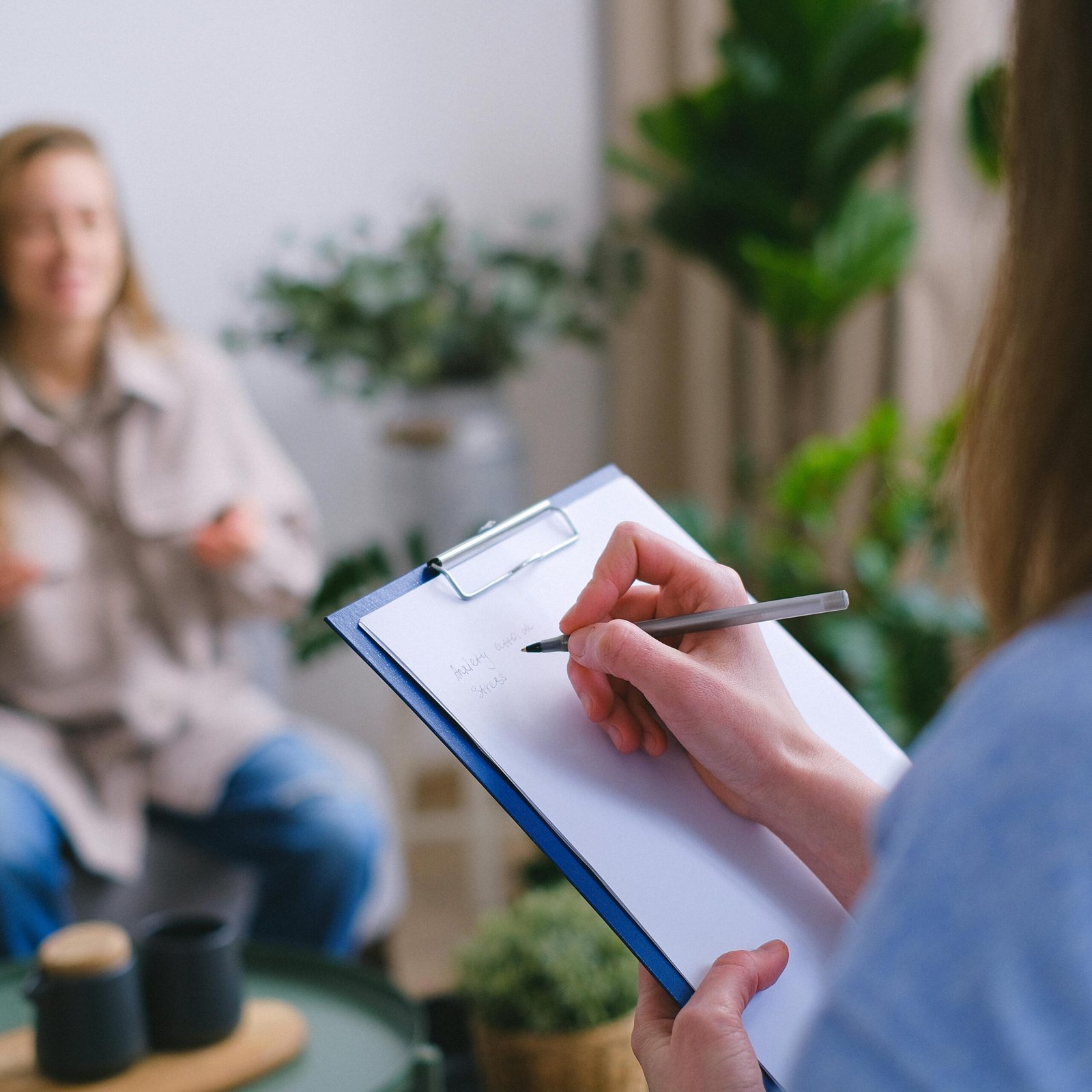 Unrecognizable professional female psychologist writing on clipboard while sitting against client on blurred background during psychotherapy session in light office