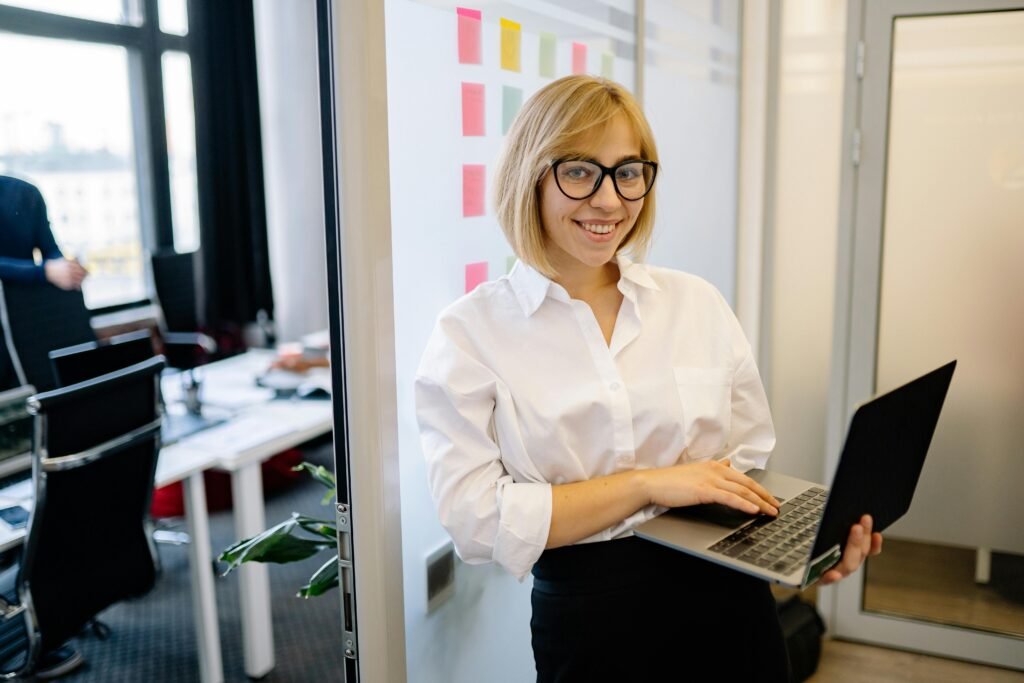 Young woman in glasses smiling confidently in a modern office setting with a laptop.